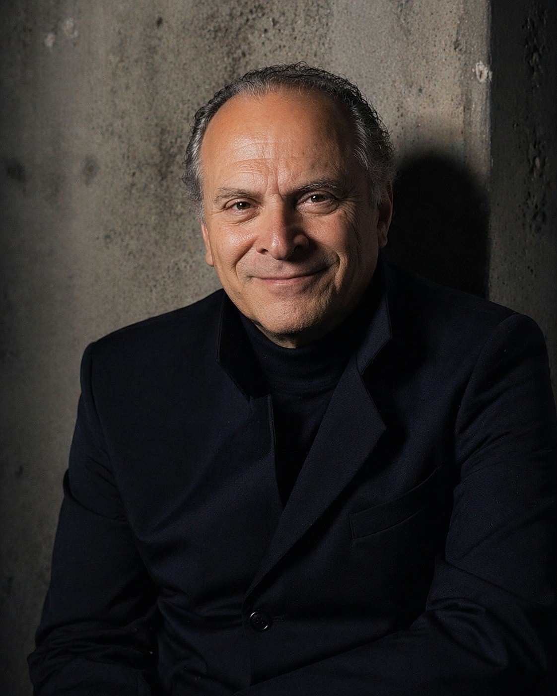 Man in black suit smiling against a textured wall.